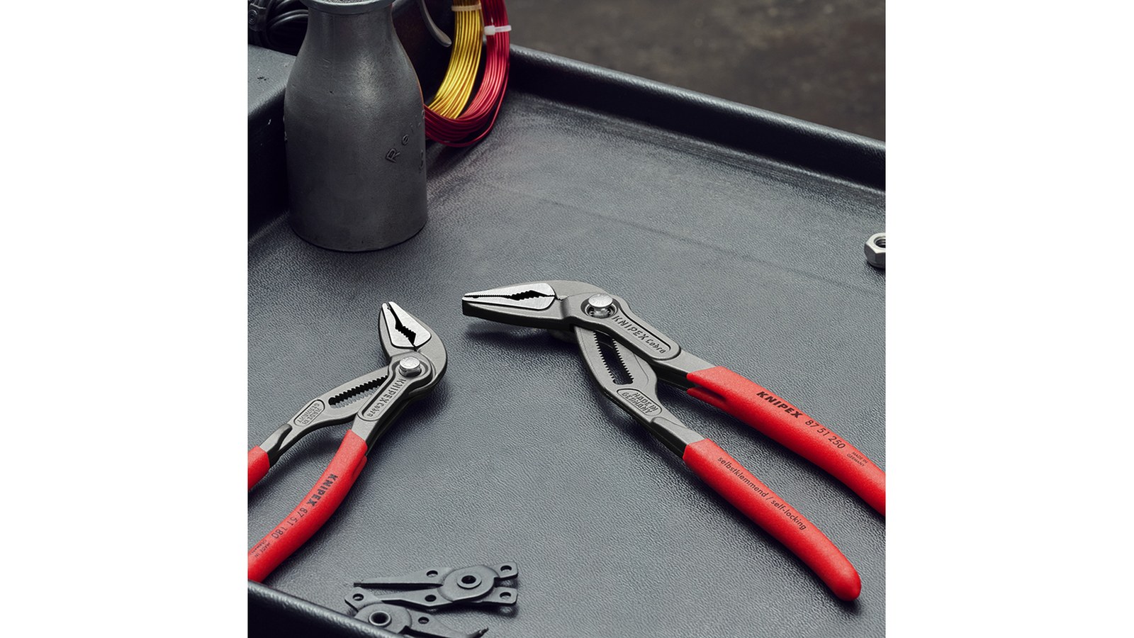 Knipex Cobra water-pump pliers with red handles lying on a dark tray beside small hardware