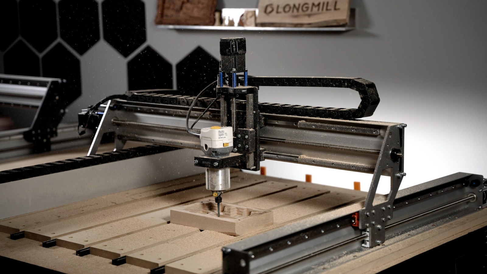 Sienci LongMill MK3 CNC router with a trim router mounted on the gantry, cutting a wooden part on a slatted table, with a LongMill sign on the wall behind