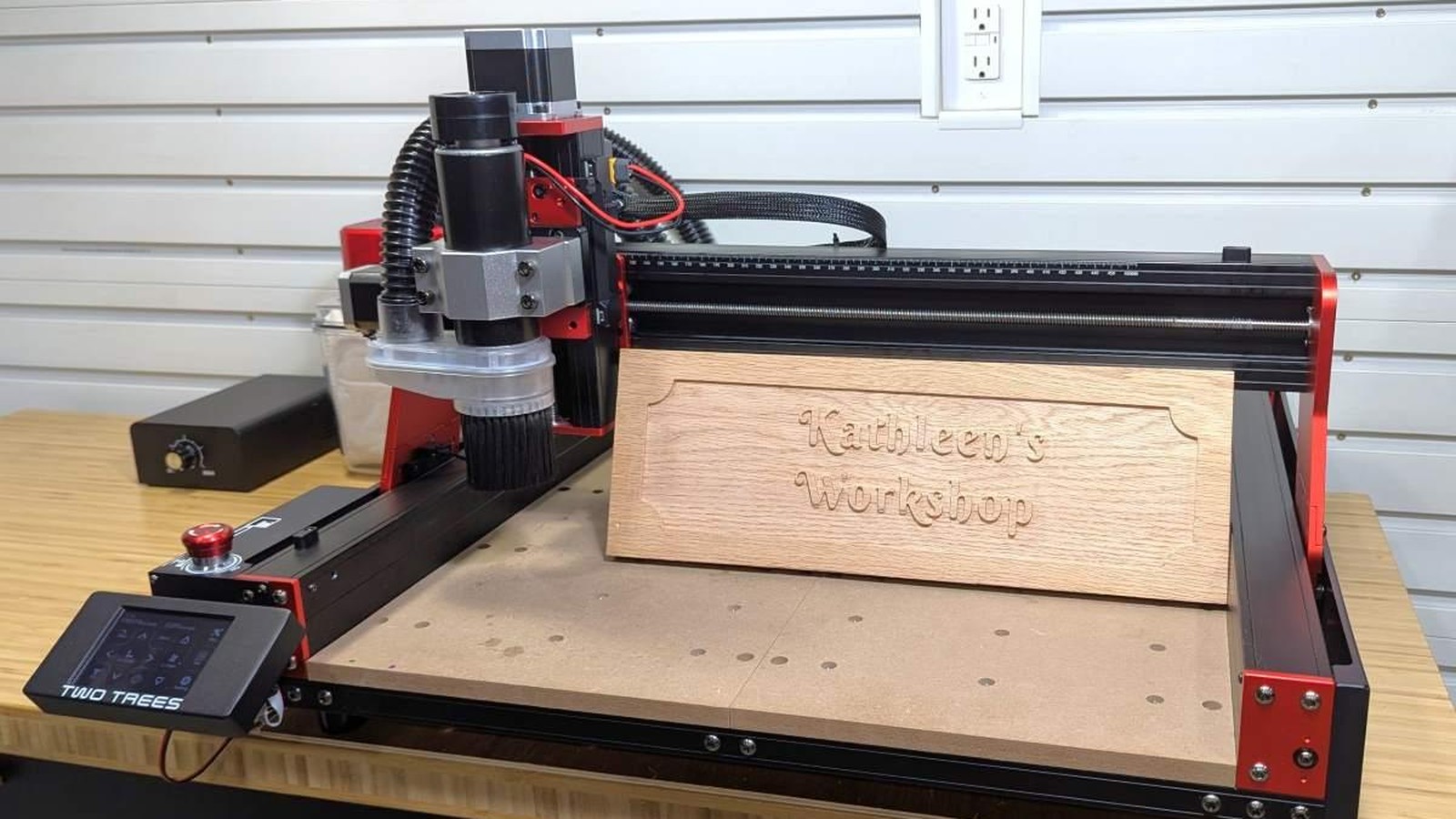 TwoTrees TTC 450 Pro CNC router with a red and black frame cutting a carved wooden workshop sign on a workbench, with its touchscreen controller at the front left