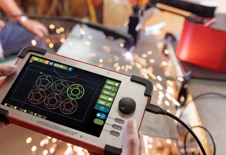 Hands holding the ArcDroid Plus controller with a touchscreen showing circle cut paths and a joystick, with plasma cutting sparks in the background