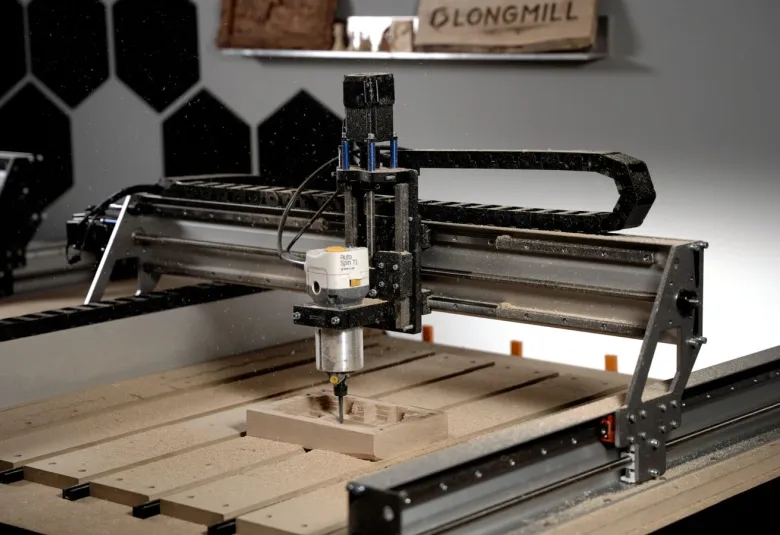Sienci LongMill MK3 CNC router with a trim router mounted on the gantry, cutting a wooden part on a slatted table, with a LongMill sign on the wall behind