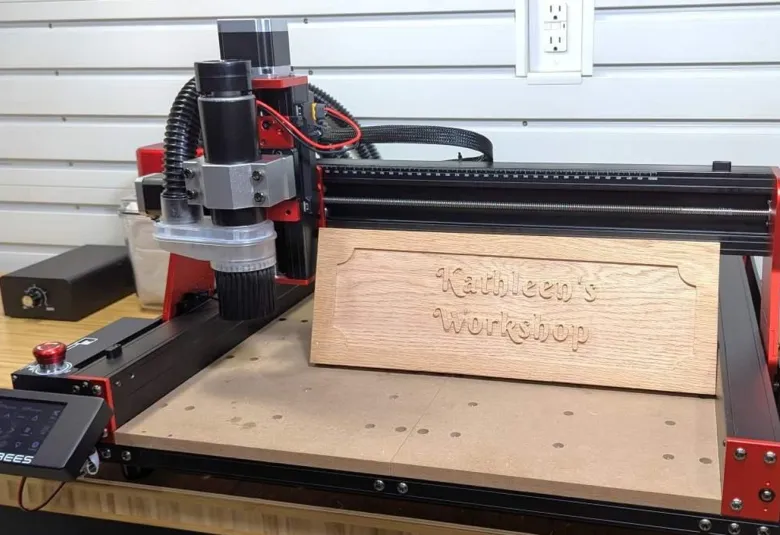 TwoTrees TTC 450 Pro CNC router with a red and black frame cutting a carved wooden workshop sign on a workbench, with its touchscreen controller at the front left