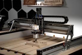 Sienci LongMill MK3 CNC router with a trim router mounted on the gantry, cutting a wooden part on a slatted table, with a LongMill sign on the wall behind