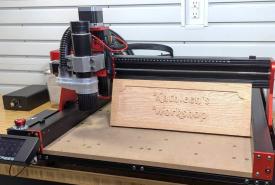 TwoTrees TTC 450 Pro CNC router with a red and black frame cutting a carved wooden workshop sign on a workbench, with its touchscreen controller at the front left
