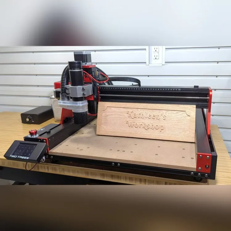 TwoTrees TTC 450 Pro CNC router with a red and black frame cutting a carved wooden workshop sign on a workbench, with its touchscreen controller at the front left
