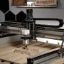 Sienci LongMill MK3 CNC router with a trim router mounted on the gantry, cutting a wooden part on a slatted table, with a LongMill sign on the wall behind