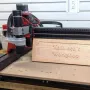 TwoTrees TTC 450 Pro CNC router with a red and black frame cutting a carved wooden workshop sign on a workbench, with its touchscreen controller at the front left